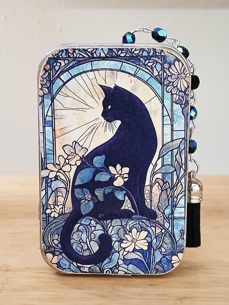 Puede incluir: Caja de baratijas rectangular decorativa con la silueta de un gato negro en un dise&ntilde;o de vidriera. La obra de arte incluye flores blancas y follaje azul sobre un fondo azul claro. Una cadena de cuentas y una borla negra est&aacute;n adjuntas.
