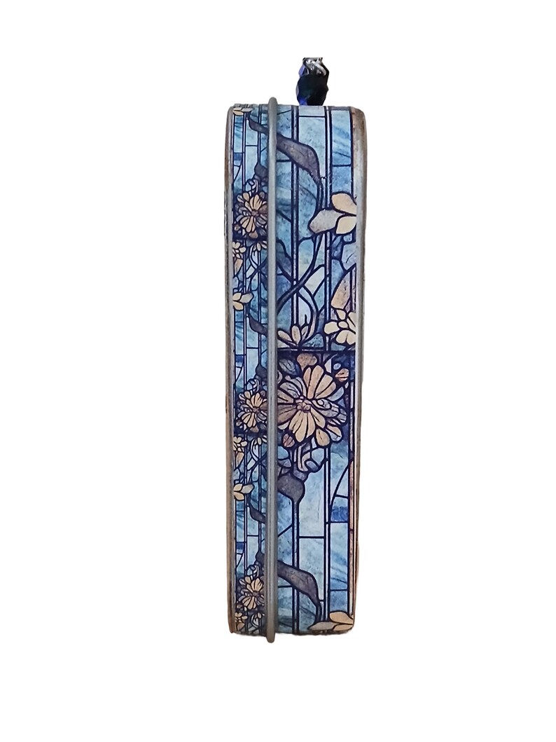 Puede incluir: Una caja decorativa rectangular con un dise&ntilde;o floral. La caja presenta un efecto de vidriera azul con flores beige y hojas verdes. Un marco de metal plateado rodea la caja, y un peque&ntilde;o elemento decorativo azul oscuro se encuentra en la parte superior.