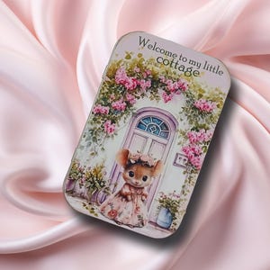 May include: A rectangular tin featuring a whimsical illustration. The design includes a mouse in a floral dress standing before a cottage door adorned with pink flowers. The text "Welcome to my little cottage" is at the top.