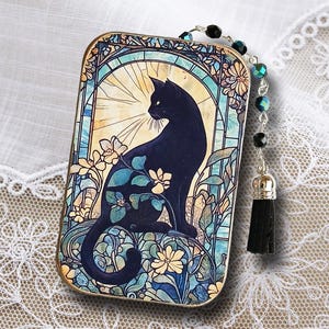 Puede incluir: Colgante rectangular con un dise&ntilde;o de gato negro al estilo vidriera. El gato est&aacute; rodeado de patrones florales azules, amarillos y verdes. Una cadena de cuentas con una borla negra est&aacute; unida al colgante.