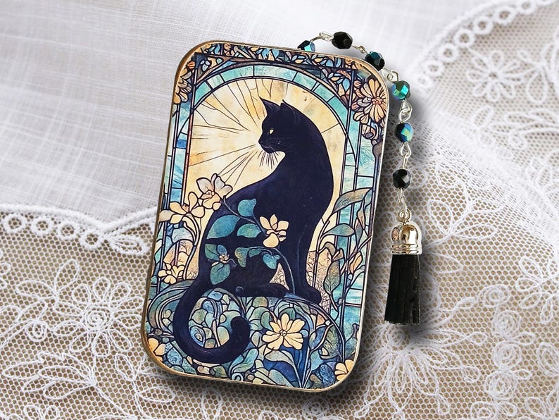 Puede incluir: Colgante rectangular con un dise&ntilde;o de gato negro al estilo vidriera. El gato est&aacute; rodeado de patrones florales azules, amarillos y verdes. Una cadena de cuentas con una borla negra est&aacute; unida al colgante.
