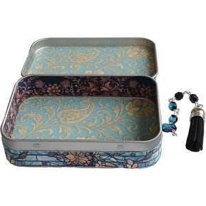 Puede incluir: Lata rectangular abierta con un interior de estampado de cachemira azul claro y dorado. El exterior tiene un dise&ntilde;o azul y floral. Un dije de cuentas con una borla negra cuelga a la derecha.