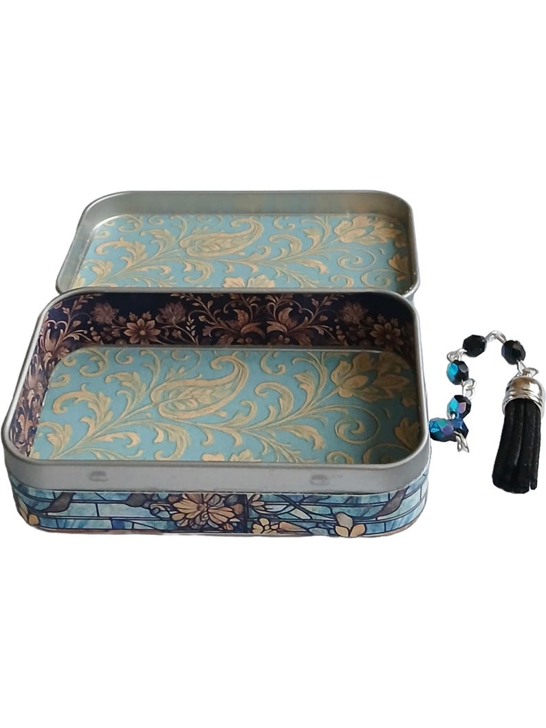 Puede incluir: Lata rectangular abierta con un interior de estampado de cachemira azul claro y dorado. El exterior tiene un dise&ntilde;o azul y floral. Un dije de cuentas con una borla negra cuelga a la derecha.