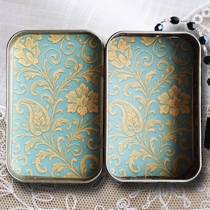 Puede incluir: Dos cajas rectangulares de metal con bisagras, con fondo azul claro y un patr&oacute;n floral dorado. Una cadena de cuentas negras con borla cuelga de una caja. Las cajas est&aacute;n abiertas, revelando el dise&ntilde;o detallado.