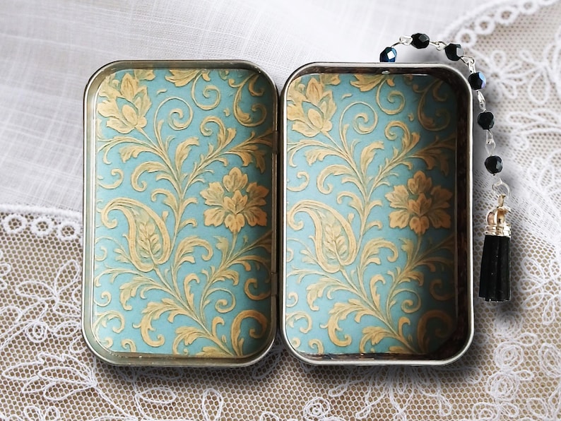 Puede incluir: Dos cajas rectangulares de metal con bisagras, con fondo azul claro y un patr&oacute;n floral dorado. Una cadena de cuentas negras con borla cuelga de una caja. Las cajas est&aacute;n abiertas, revelando el dise&ntilde;o detallado.