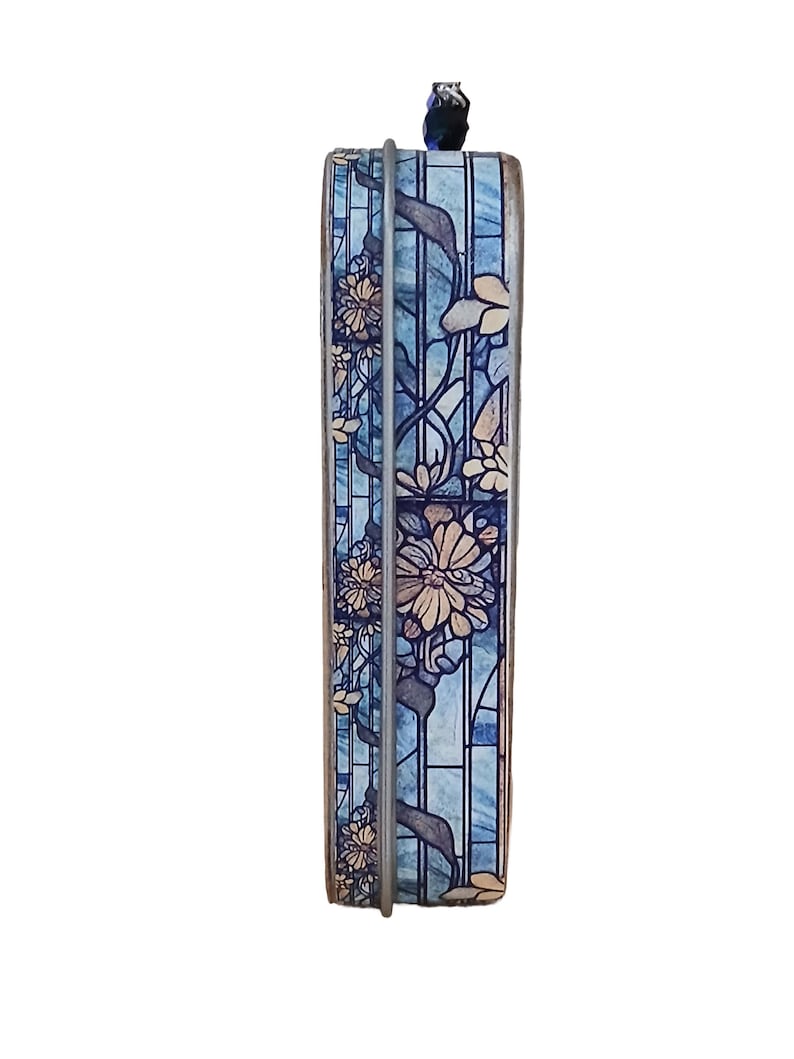 Puede incluir: Una caja decorativa rectangular con un dise&ntilde;o floral. La caja presenta un efecto de vidriera azul con flores beige y hojas verdes. Un marco de metal plateado rodea la caja, y un peque&ntilde;o elemento decorativo azul oscuro se encuentra en la parte superior.