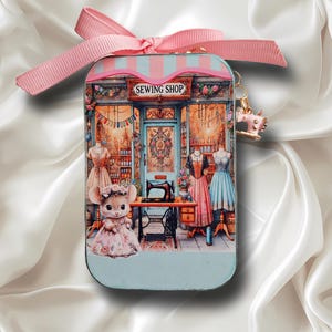 May include: A decorative tin featuring a whimsical sewing shop scene. The shop has a blue facade with a pink awning and the text "SEWING SHOP". Inside, a mouse in a dress stands near a sewing machine. A pink ribbon tops the tin.