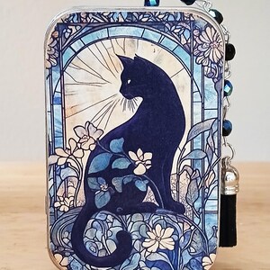 Puede incluir: Caja de baratijas rectangular decorativa con la silueta de un gato negro en un dise&ntilde;o de vidriera. La obra de arte incluye flores blancas y follaje azul sobre un fondo azul claro. Una cadena de cuentas y una borla negra est&aacute;n adjuntas.