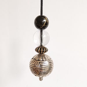 May include: A black cord with three beads hanging from it. The top bead is black with gold accents. The middle bead is clear glass. The bottom bead is silver with a geometric pattern.