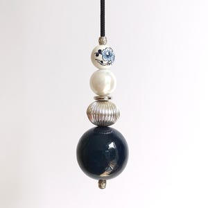 May include: A decorative pull cord featuring a vertical arrangement of beads suspended from a black string. The beads include a small white ceramic sphere with a blue floral pattern, a smooth white sphere, a ribbed silver-toned bead, and a large glossy black sphere.
