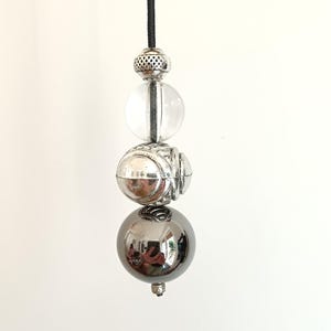 May include: A vertical pull cord featuring a series of decorative beads. The arrangement includes a small textured silver bead, a clear spherical bead, a larger oval silver bead with etched details, and a smooth, reflective dark metallic sphere.