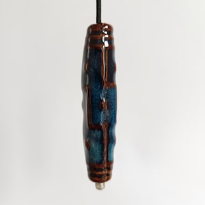 May include: A ceramic light pull with a dark blue and brown glaze. The cylindrical pull has a textured surface and is suspended by a black cord. The bottom has a small silver metal cap.