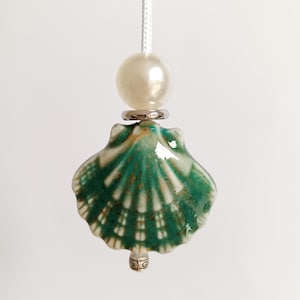 May include: A ceramic seashell light pull with a pearl bead and silver-tone accents. The seashell is teal and white, with a glossy finish. The light pull hangs from a white cord, adding a touch of coastal charm to any room.