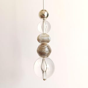 May include: A decorative hanging ornament with clear and silver beads. The beads include a silver sphere, two clear spheres, and two textured silver beads. The ornament hangs against a white backdrop.