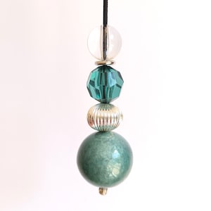 May include: A decorative pull cord featuring a vertical arrangement of beads suspended from a black string. The beads include a clear sphere, a faceted teal crystal, a ribbed metallic silver bead, and a large, smooth, mottled green sphere.