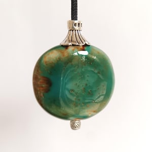 May include: A round green and brown ceramic light pull with silver accents. The pull is attached to a black cord.