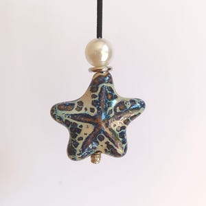 May include: A star-shaped pendant featuring a glossy, mottled glaze in shades of blue, brown, and cream. The pendant is suspended from a black cord and accented with a single white pearl bead and a small silver-toned metal spacer at the top.