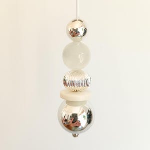 May include: A vertical light pull cord featuring a sequence of five decorative beads. The arrangement includes a smooth silver sphere, a translucent white sphere, a ribbed silver bead, a flat white disc, and a large reflective silver sphere.