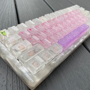 May include: A custom keyboard with clear, glittery keycaps and a pearlescent case. The keys have pink lettering and a green alien-shaped key. The keyboard features a gradient of pink and purple keys.
