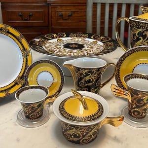 May include: A collection of luxury tableware featuring a yellow and black ornate design. Includes a large plate, a serving platter, a teapot, saucers, cups, a creamer, and a covered dish. The set has a gold trim.