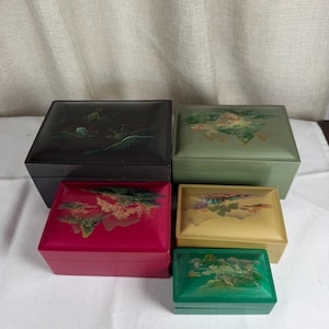 May include: A collection of five rectangular lidded boxes in black, sage green, magenta, gold, and emerald green. Each box features a painted landscape illustration on the lid, depicting mountains, trees, and foliage in various colors.