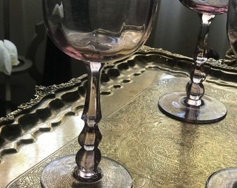 May include: Four clear, pink-tinted stemmed wine glasses on a gold, ornate tray. The glasses have a wide bowl and a delicate stem.
