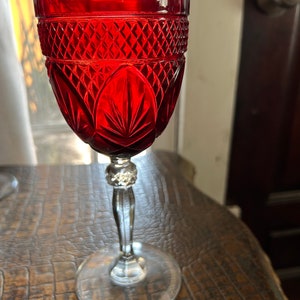 May include: A red glass goblet with a diamond pattern and a clear stem. The goblet is sitting on a brown surface.