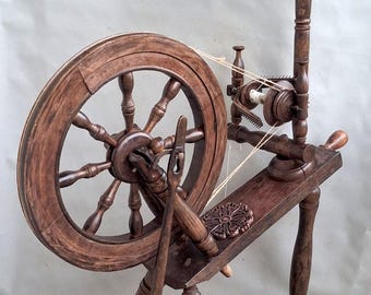 May include: A dark brown wooden spinning wheel featuring a large circular wheel with multiple spokes, a treadle mechanism, a flyer assembly with a bobbin, and a vertical distaff. The structure is supported by three turned legs and includes a decorative carved floral detail on the main horizontal frame.