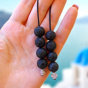 May include: Black lava rock bead earrings with silver cross charms. The earrings are strung on black cords and held in a hand. The background shows blurred white buildings with blue domes, suggesting a coastal or island setting.