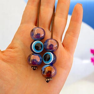 Puede incluir: Primer plano de varias cuentas de vidrio azules y moradas con un diseño de ojo, ensartadas en cordones marrones. Las cuentas tienen un brillo metálico y están acentuadas con detalles blancos y negros. Es un artículo decorativo.