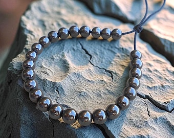 May include: A dark gray beaded bracelet with adjustable cord closure. The bracelet features multiple polished, spherical beads. The bracelet is displayed on a gray stone surface, with a blurred background.