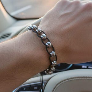 May include: A silver bead bracelet with black cord is worn on a wrist. The bracelet is made of multiple silver beads connected by black cord. The person is driving a car, with the steering wheel in view.