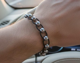 May include: A silver bead bracelet with black cord is worn on a wrist. The bracelet is made of multiple silver beads connected by black cord. The person is driving a car, with the steering wheel in view.