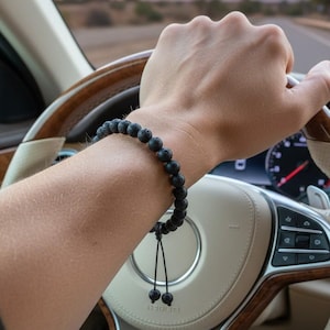 Pode incluir: Uma pulseira de contas de pedra de lava preta é usada no pulso de uma pessoa que dirige um carro. O volante é de madeira e couro creme. O fundo mostra uma paisagem desértica e um pôr do sol.