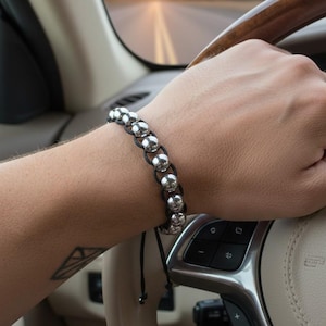 Puede incluir: Una pulsera de cuentas plateadas con cordón negro se lleva en la muñeca. La pulsera presenta múltiples esferas plateadas ensartadas juntas. El fondo muestra el interior de un coche con un volante y una carretera al atardecer.