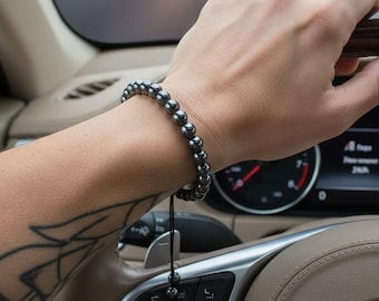 May include: A dark gray beaded bracelet is worn on a person's wrist, with the person's hand on a car's steering wheel. The bracelet is made of round, polished beads. The car's interior is beige, and the background shows a road and a sunset.