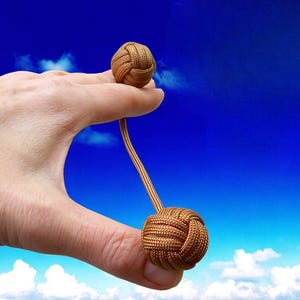 May include: A brown decorative knot, held between fingers, with a smaller knot attached by a cord. The knots are made of woven material. The background is a bright blue sky with white clouds.