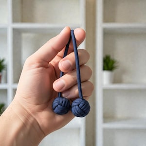 May include: Two navy blue decorative monkey fist knot balls connected by a navy blue cord. The balls are held in a hand against a blurred white background.