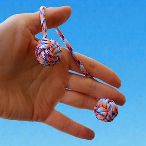 May include: Two decorative knotted balls made of orange, blue, and purple cord are held in a hand against a blue background. The balls are connected by the same cord, creating a unique design. The balls are approximately 3.8 cm in diameter.