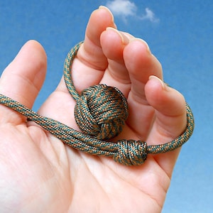 May include: A decorative knot made of green and brown cord. The knot is round and has a smaller knot attached to a cord loop. The background is a blue sky.