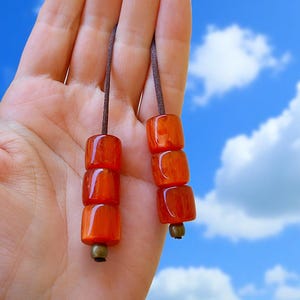 May include: Two strands of orange-red beads, each with three square beads, suspended from a brown cord. The beads have a glossy finish and are accented with small, antique-style metal beads. The background is a bright blue sky with fluffy white clouds.