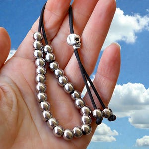May include: A silver beaded bracelet with a skull charm. The bracelet features multiple small, round, silver beads strung on a black cord. A small silver skull charm is centered on the cord. The bracelet is held in a hand against a blue sky background.