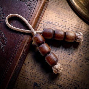 Puede incluir: Un rosario de cuentas de madera hecho a mano con un cordón beige y extremos anudados. Las cuentas son de color marrón oscuro y tienen una forma lisa y redondeada. El rosario descansa sobre un libro encuadernado en cuero marrón.