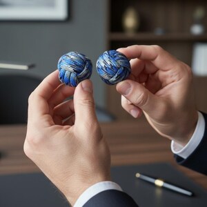 May include: Two blue and gray knot cufflinks held between two hands. The cufflinks are round and made of woven material. The background is blurred, with a pen visible on a dark surface.