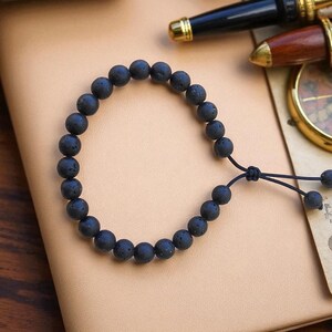 May include: Black lava stone bead bracelet with an adjustable cord closure. The bracelet features round, dark gray beads. The bracelet is displayed on a light brown surface, with a pen and compass in the background.