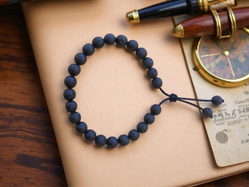 May include: Black lava stone bead bracelet with an adjustable cord closure. The bracelet features round, dark gray beads. The bracelet is displayed on a light brown surface, with a pen and compass in the background.
