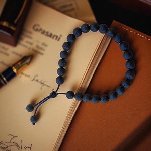 May include: A dark blue lava stone bead bracelet with a sliding knot closure. The bracelet is resting on a notebook and a leather-bound journal. A pen and ink bottle are also visible.