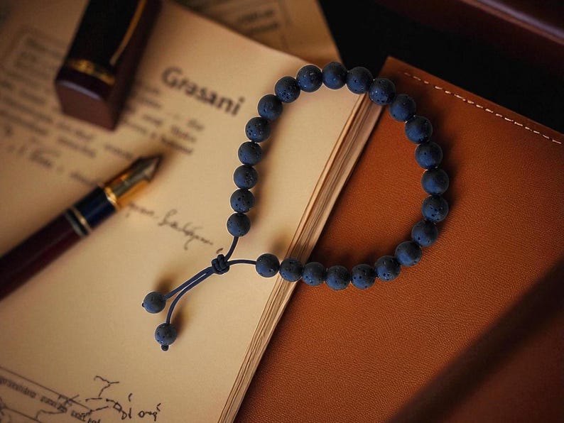 May include: A dark blue lava stone bead bracelet with a sliding knot closure. The bracelet is resting on a notebook and a leather-bound journal. A pen and ink bottle are also visible.