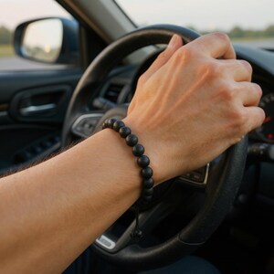 May include: A black beaded bracelet with a sliding knot closure. The bracelet is worn on a wrist, with a car steering wheel in the background. The beads appear to be approximately 0.4 inches in diameter.