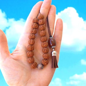 Puede incluir: Un collar de cuentas mala marrón con borla y detalles decorativos. Las cuentas son de color natural y la borla es marrón. El collar se muestra sobre un fondo de cielo azul.